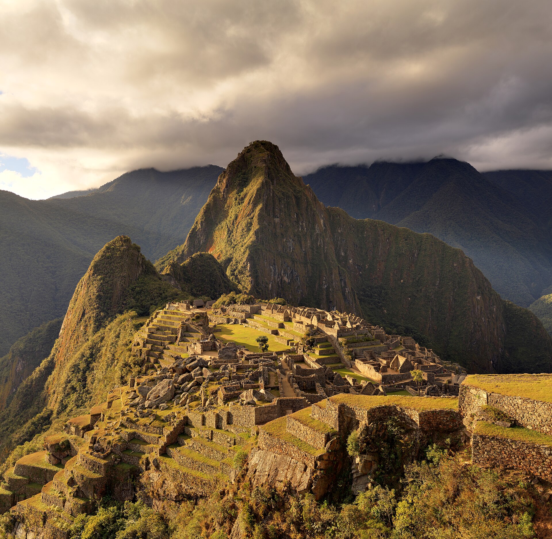 Natural golden evening light across Machu Picchu's citadel and Huayna Picchu, with clouds above the surrounding Andes